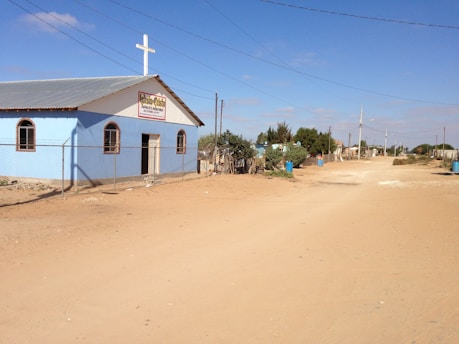 A small blue church with a cross on top is situated alongside a quiet dirt road under a clear blue sky. Surrounding the church is a chain-link fence, and nearby are some bushes and blue barrels. Electricity lines run parallel to the road leading to a small settlement visible in the distance.