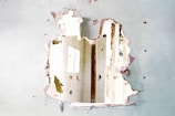 A large hole is visible in a wall with peeling paint and crumbling plaster, revealing a view of a bright interior with doors and possibly windows. The wall appears to be in a state of disrepair.