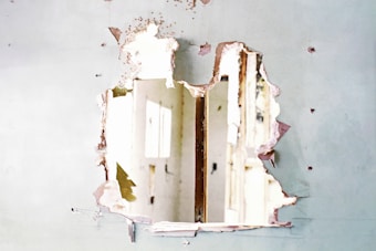 A large hole is visible in a wall with peeling paint and crumbling plaster, revealing a view of a bright interior with doors and possibly windows. The wall appears to be in a state of disrepair.