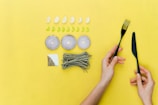 A yellow background features a neatly arranged set of food ingredients and utensils. There are five cloves of garlic and eight slices of green pepper placed in rows. Three onion rings are arranged in a line, and a bunch of green pasta is placed next to a square section of salt and pepper. A pair of hands holds a black fork and knife on the right side of the image.