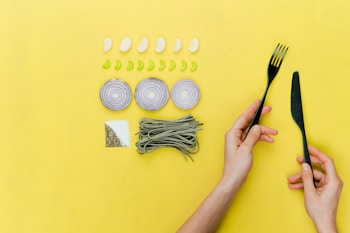 A yellow background features a neatly arranged set of food ingredients and utensils. There are five cloves of garlic and eight slices of green pepper placed in rows. Three onion rings are arranged in a line, and a bunch of green pasta is placed next to a square section of salt and pepper. A pair of hands holds a black fork and knife on the right side of the image.