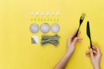 A yellow background features a neatly arranged set of food ingredients and utensils. There are five cloves of garlic and eight slices of green pepper placed in rows. Three onion rings are arranged in a line, and a bunch of green pasta is placed next to a square section of salt and pepper. A pair of hands holds a black fork and knife on the right side of the image.