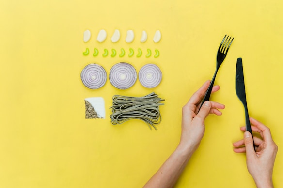 A yellow background features a neatly arranged set of food ingredients and utensils. There are five cloves of garlic and eight slices of green pepper placed in rows. Three onion rings are arranged in a line, and a bunch of green pasta is placed next to a square section of salt and pepper. A pair of hands holds a black fork and knife on the right side of the image.