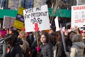 A large group of people participating in a protest or rally, holding various signs with messages related to social or political causes. The central sign reads 'Vote me for President 2050' with a red arrow pointing downward. Many participants are wearing winter clothing, indicating a cold weather setting.