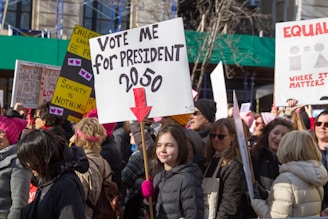 A large group of people participating in a protest or rally, holding various signs with messages related to social or political causes. The central sign reads 'Vote me for President 2050' with a red arrow pointing downward. Many participants are wearing winter clothing, indicating a cold weather setting.