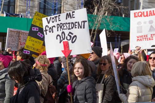 A large group of people participating in a protest or rally, holding various signs with messages related to social or political causes. The central sign reads 'Vote me for President 2050' with a red arrow pointing downward. Many participants are wearing winter clothing, indicating a cold weather setting.