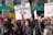 A large group of people participating in a protest or rally, holding various signs with messages related to social or political causes. The central sign reads 'Vote me for President 2050' with a red arrow pointing downward. Many participants are wearing winter clothing, indicating a cold weather setting.