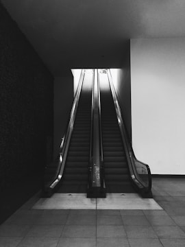 A pair of escalators rise side by side in a dimly lit indoor setting. The surroundings are minimalistic with smooth walls, and the tiles on the floor create a sense of uniformity. The escalators lead to a bright opening above, suggesting access to another level or area.