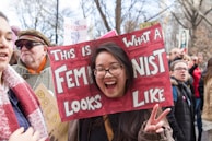 A person is smiling and holding a sign with a cutout frame spelling 'This is what a feminist looks like' during a public gathering. The background shows a crowd of people, some holding signs in a possibly protest or rally setting.