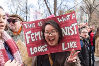A person is smiling and holding a sign with a cutout frame spelling 'This is what a feminist looks like' during a public gathering. The background shows a crowd of people, some holding signs in a possibly protest or rally setting.