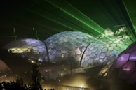 A series of large, interconnected geodesic domes illuminated by green and purple lights at night. The structures have a honeycomb pattern and extend across a misty background. Green laser beams project across the sky above, giving a futuristic ambiance.