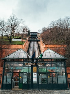 A glass-fronted funicular station with the words 'BUDAVARI SIKLO' on the entrance is positioned at the base of a steep track. The funicular ascends between brick walls, and the area is surrounded by leafless trees, suggesting a winter setting. Flags are visible above the entrance.