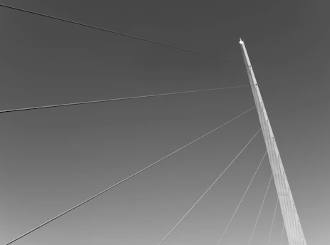 A stark, high-contrast editorial photo of a lone figure standing beneath a towering network of cables and data points, symbolizing the unseen systems shaping society.