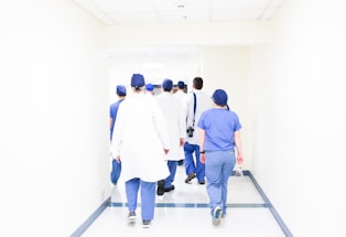 A group of medical professionals in scrubs and lab coats walks down a bright hospital corridor. They appear to be engaged in conversation, heading towards a destination within the hospital. The environment is clean and well-lit, emphasizing a clinical setting.
