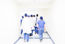 A group of medical professionals in scrubs and lab coats walks down a bright hospital corridor. They appear to be engaged in conversation, heading towards a destination within the hospital. The environment is clean and well-lit, emphasizing a clinical setting.