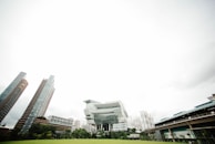 A modern urban landscape features several tall buildings with contemporary architectural designs. In the foreground, a large green lawn provides a natural contrast to the cityscape. The sky is overcast, with a vast expanse of white cloud cover.