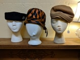 Three mannequin heads showcase different vintage hats on a wooden shelf. The head on the left features a black beret adorned with a small embellishment. The center head is crowned with a striped hat in shades of brown and black, tied with a ribbon. The right head dons a beige hat with layered folds. Two of the heads have tags hanging from them.