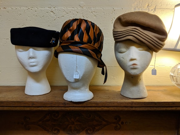 Three mannequin heads showcase different vintage hats on a wooden shelf. The head on the left features a black beret adorned with a small embellishment. The center head is crowned with a striped hat in shades of brown and black, tied with a ribbon. The right head dons a beige hat with layered folds. Two of the heads have tags hanging from them.