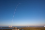 A rocket launches into a clear blue sky, leaving a thin white trail arching dramatically upwards. The launch site is surrounded by water and greenery, with the exhaust plume visible near the ground.