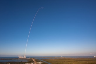 A rocket launches into a clear blue sky, leaving a thin white trail arching dramatically upwards. The launch site is surrounded by water and greenery, with the exhaust plume visible near the ground.