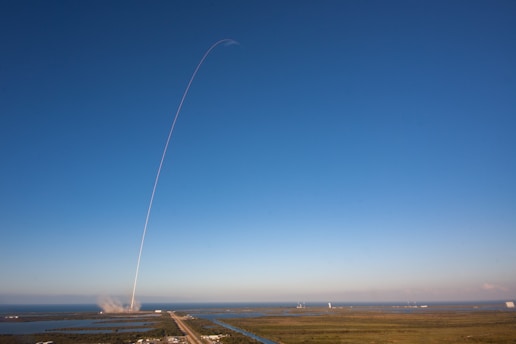 A rocket launches into a clear blue sky, leaving a thin white trail arching dramatically upwards. The launch site is surrounded by water and greenery, with the exhaust plume visible near the ground.