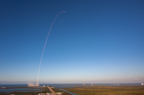 A rocket launches into a clear blue sky, leaving a thin white trail arching dramatically upwards. The launch site is surrounded by water and greenery, with the exhaust plume visible near the ground.