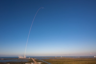 A rocket launches into a clear blue sky, leaving a thin white trail arching dramatically upwards. The launch site is surrounded by water and greenery, with the exhaust plume visible near the ground.