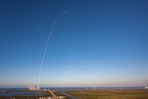 A rocket launches into a clear blue sky, leaving a thin white trail arching dramatically upwards. The launch site is surrounded by water and greenery, with the exhaust plume visible near the ground.