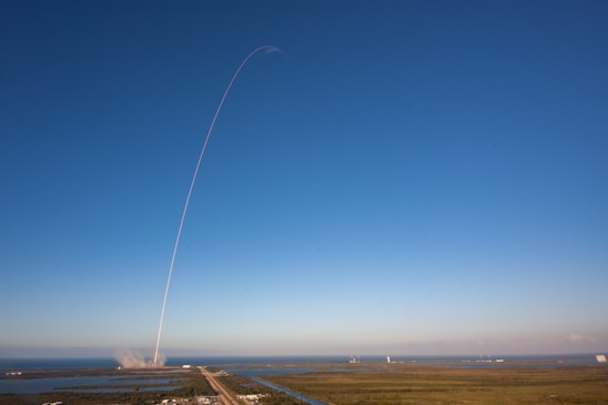 A rocket launches into a clear blue sky, leaving a thin white trail arching dramatically upwards. The launch site is surrounded by water and greenery, with the exhaust plume visible near the ground.