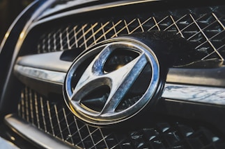 A close-up view of a car grille featuring a prominent metallic emblem of the Hyundai logo. The grille has a mesh pattern, and the lighting highlights the chrome accents.