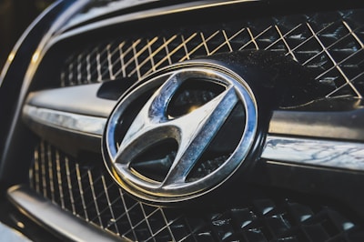 A close-up view of a car grille featuring a prominent metallic emblem of the Hyundai logo. The grille has a mesh pattern, and the lighting highlights the chrome accents.