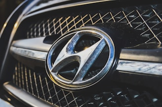 A close-up view of a car grille featuring a prominent metallic emblem of the Hyundai logo. The grille has a mesh pattern, and the lighting highlights the chrome accents.