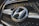 A close-up view of a car grille featuring a prominent metallic emblem of the Hyundai logo. The grille has a mesh pattern, and the lighting highlights the chrome accents.