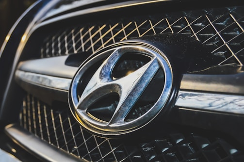 A close-up view of a car grille featuring a prominent metallic emblem of the Hyundai logo. The grille has a mesh pattern, and the lighting highlights the chrome accents.