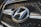 A close-up view of a car grille featuring a prominent metallic emblem of the Hyundai logo. The grille has a mesh pattern, and the lighting highlights the chrome accents.