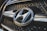 A close-up view of a car grille featuring a prominent metallic emblem of the Hyundai logo. The grille has a mesh pattern, and the lighting highlights the chrome accents.