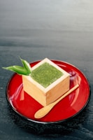 A cube-shaped wooden container holds a green powder, likely matcha, topped with a leaf garnish. It is placed on a glossy red plate alongside a small wooden spoon.
