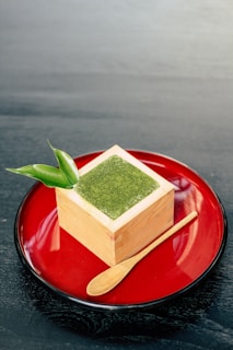 A cube-shaped wooden container holds a green powder, likely matcha, topped with a leaf garnish. It is placed on a glossy red plate alongside a small wooden spoon.