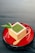 A cube-shaped wooden container holds a green powder, likely matcha, topped with a leaf garnish. It is placed on a glossy red plate alongside a small wooden spoon.