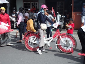 A group of people gathered outdoors, possibly participating in a parade or cultural event. A woman stands prominently in the center, holding a megaphone and wearing a cap and casual clothing. She is next to a bicycle decorated with red and white streamers. Several other individuals, including children, are dressed in colorful traditional attire and there are onlookers in the background.