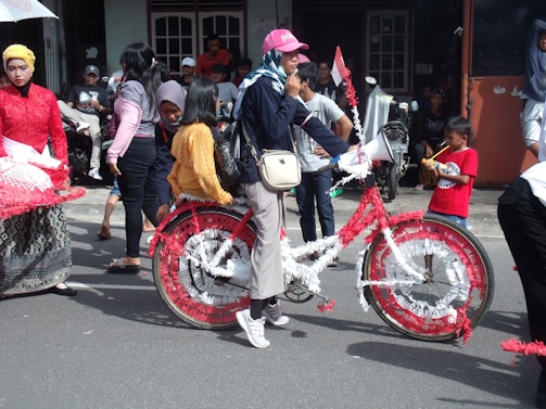A group of people gathered outdoors, possibly participating in a parade or cultural event. A woman stands prominently in the center, holding a megaphone and wearing a cap and casual clothing. She is next to a bicycle decorated with red and white streamers. Several other individuals, including children, are dressed in colorful traditional attire and there are onlookers in the background.