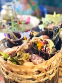 An assortment of food items presented in a woven basket, featuring grilled meat, eggs, fresh greens, and colorful flowers. The background is softly blurred, suggesting a vibrant setting with a mix of traditional and festive elements.