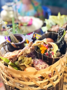 An assortment of food items presented in a woven basket, featuring grilled meat, eggs, fresh greens, and colorful flowers. The background is softly blurred, suggesting a vibrant setting with a mix of traditional and festive elements.