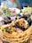 An assortment of food items presented in a woven basket, featuring grilled meat, eggs, fresh greens, and colorful flowers. The background is softly blurred, suggesting a vibrant setting with a mix of traditional and festive elements.
