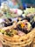 An assortment of food items presented in a woven basket, featuring grilled meat, eggs, fresh greens, and colorful flowers. The background is softly blurred, suggesting a vibrant setting with a mix of traditional and festive elements.