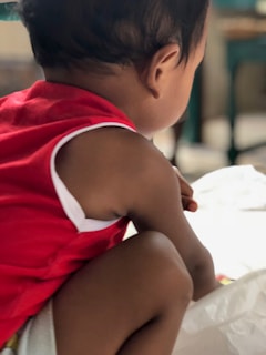 A child wearing a red sleeveless shirt is crouched down, seen from behind. The environment is softly lit with some blurred background elements, possibly furniture or indoor decor.