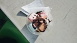 A round plastic container filled with colorful marshmallows and chocolate-covered pieces, topped with chocolate sprinkles. A small plastic spoon is placed inside the container, which is resting on a white paper napkin on a scratched, light-colored surface.