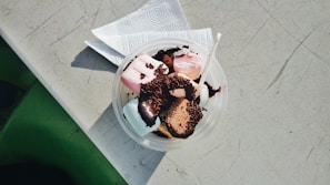 A round plastic container filled with colorful marshmallows and chocolate-covered pieces, topped with chocolate sprinkles. A small plastic spoon is placed inside the container, which is resting on a white paper napkin on a scratched, light-colored surface.
