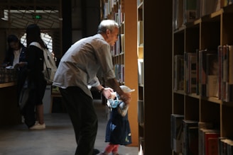 A man and a child are standing in a library aisle, with the man gently holding the child's hands. Bookshelves filled with books line both sides of the aisle. The scene is softly illuminated by natural light filtering through the windows, creating a warm and tranquil atmosphere. In the background, two more people are browsing the shelves.