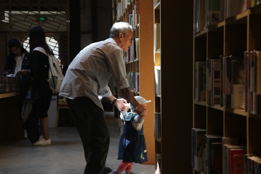 A man and a child are standing in a library aisle, with the man gently holding the child's hands. Bookshelves filled with books line both sides of the aisle. The scene is softly illuminated by natural light filtering through the windows, creating a warm and tranquil atmosphere. In the background, two more people are browsing the shelves.